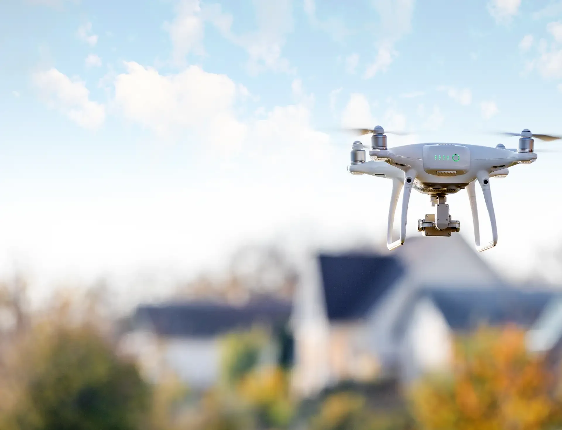 A white quadcopter drone hovering in the air with blurred houses and trees in the background